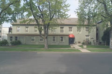 Two-story green senior residence with red entrance awning, trees, and a sidewalk.