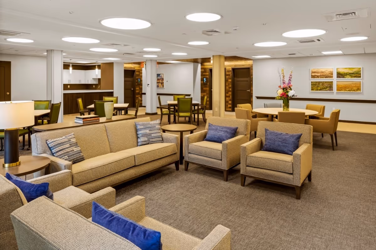 A spacious and well-lit common area in a senior living facility featuring beige sofas and armchairs with blue and patterned cushions, round tables with green and beige chairs, a side table with a lamp, and a vase of flowers on one table. The walls are decorated with framed landscape artwork, and the ceiling has multiple circular recessed lights.