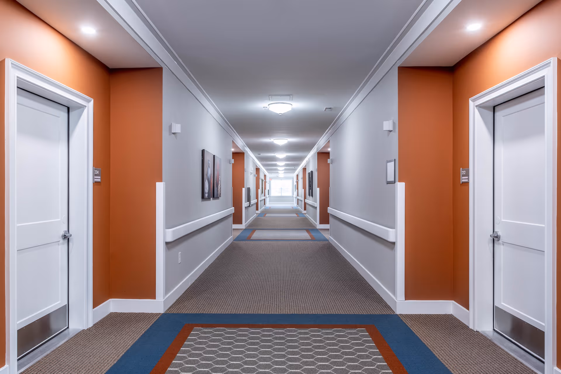 A long, well-lit interior hallway with doors on both sides, handrails, and patterned carpet.