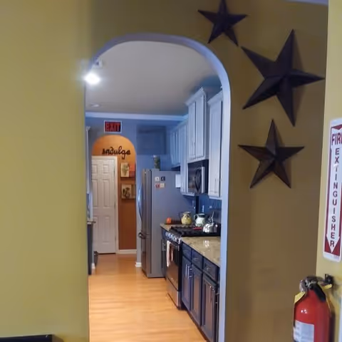 View through an arched doorway into a kitchen with white upper cabinets and dark lower cabinets, a stainless steel refrigerator, stove, and microwave. The kitchen has a granite countertop and wooden floor. On the right wall outside the kitchen are three decorative star-shaped wall hangings and a fire extinguisher mounted below a fire extinguisher sign.