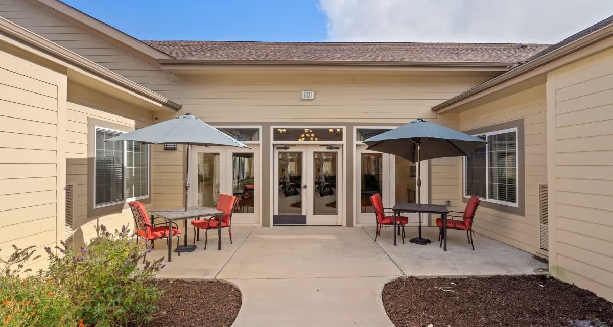 Outdoor patio area at Poet's Walk San Antonio, A Memory Care Community, featuring two tables with red cushioned chairs and large umbrellas, surrounded by beige building walls and windows, with a double glass door entrance in the center.