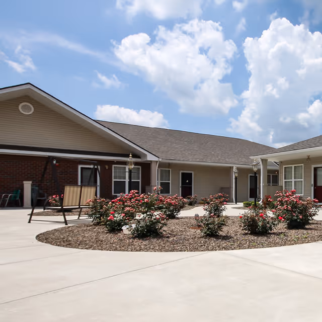 Outdoor courtyard area of a senior living facility with a circular flower bed filled with blooming red and pink roses, surrounded by a concrete walkway. The building has beige siding and brick accents with several doors and windows visible. There is a swing bench on the left side and lamp posts near the flower bed under a partly cloudy blue sky.