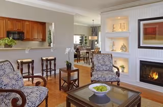 A cozy living room featuring two patterned armchairs, a glass coffee table with a bowl of green apples, a fireplace with built-in shelves displaying decorative items, and a view into a kitchen area with wooden cabinets and bar stools.