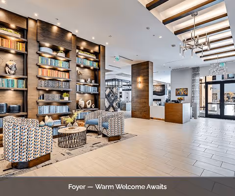 A spacious and modern foyer area with patterned armchairs arranged around a circular coffee table on a rug. Behind the seating area is a large built-in wooden bookshelf filled with books and decorative items. The space features light-colored tile flooring, a wooden accent wall, and a reception desk near the entrance with glass doors allowing natural light to enter. The ceiling has recessed lighting and a contemporary chandelier.