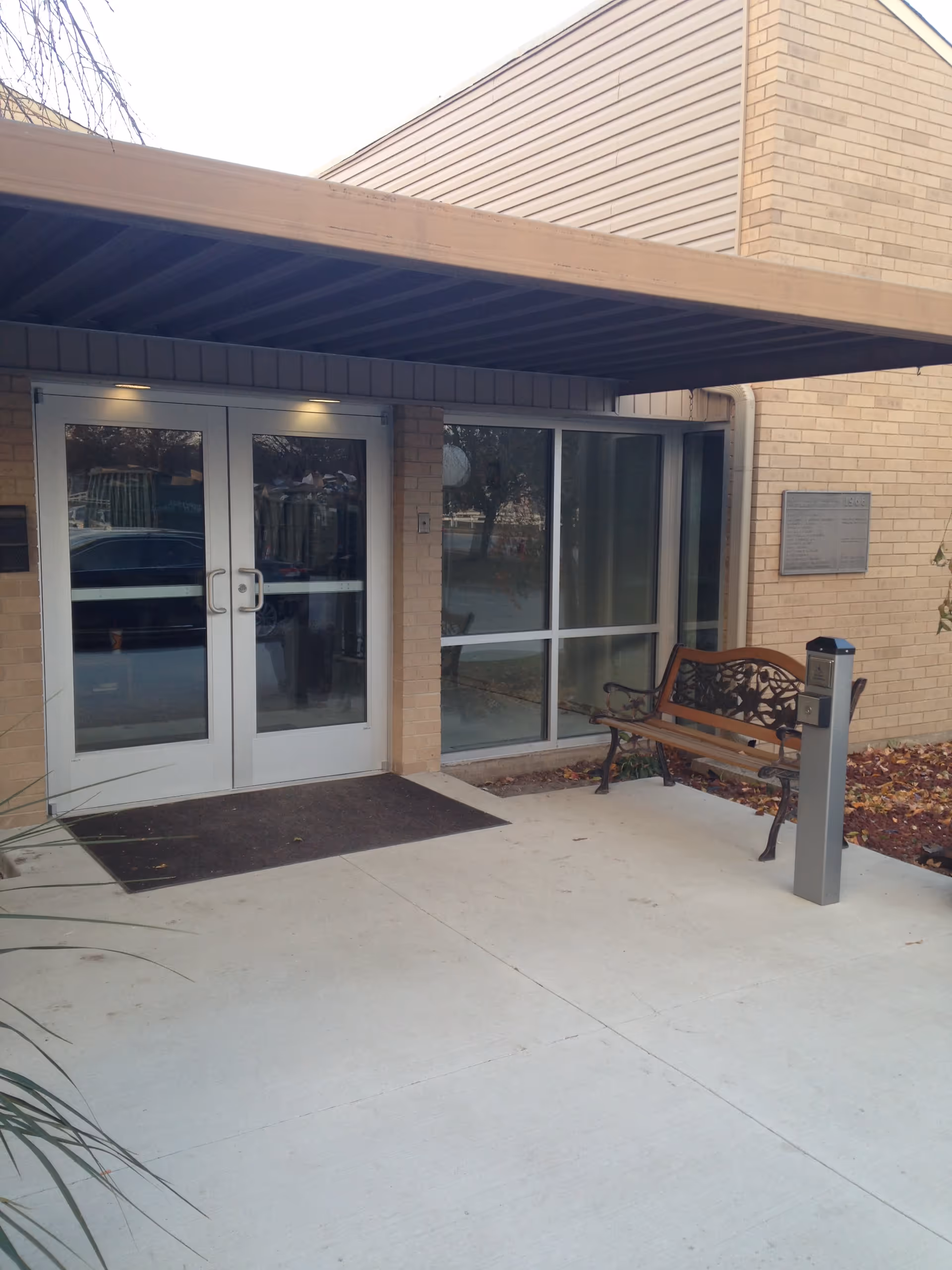 Entrance to a building with double glass doors and a large window to the right. There is a brown bench with decorative metal armrests and a small post with an intercom or access control device near the bench. The building exterior is made of light-colored bricks, and there is a covered overhang above the entrance.
