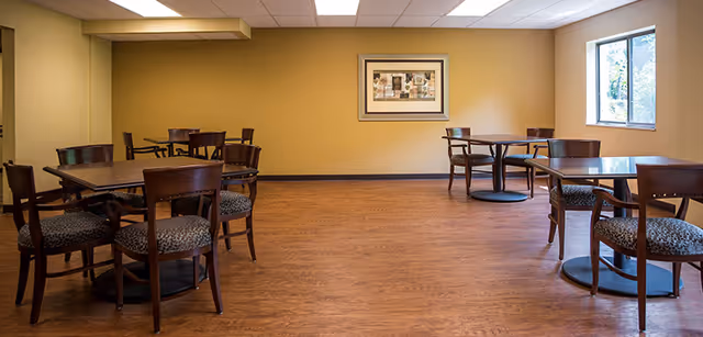 A dining room with several wooden tables and chairs arranged on a wood floor. The walls are painted a light beige color, with a framed artwork hanging on the far wall. A window on the right side lets in natural light.