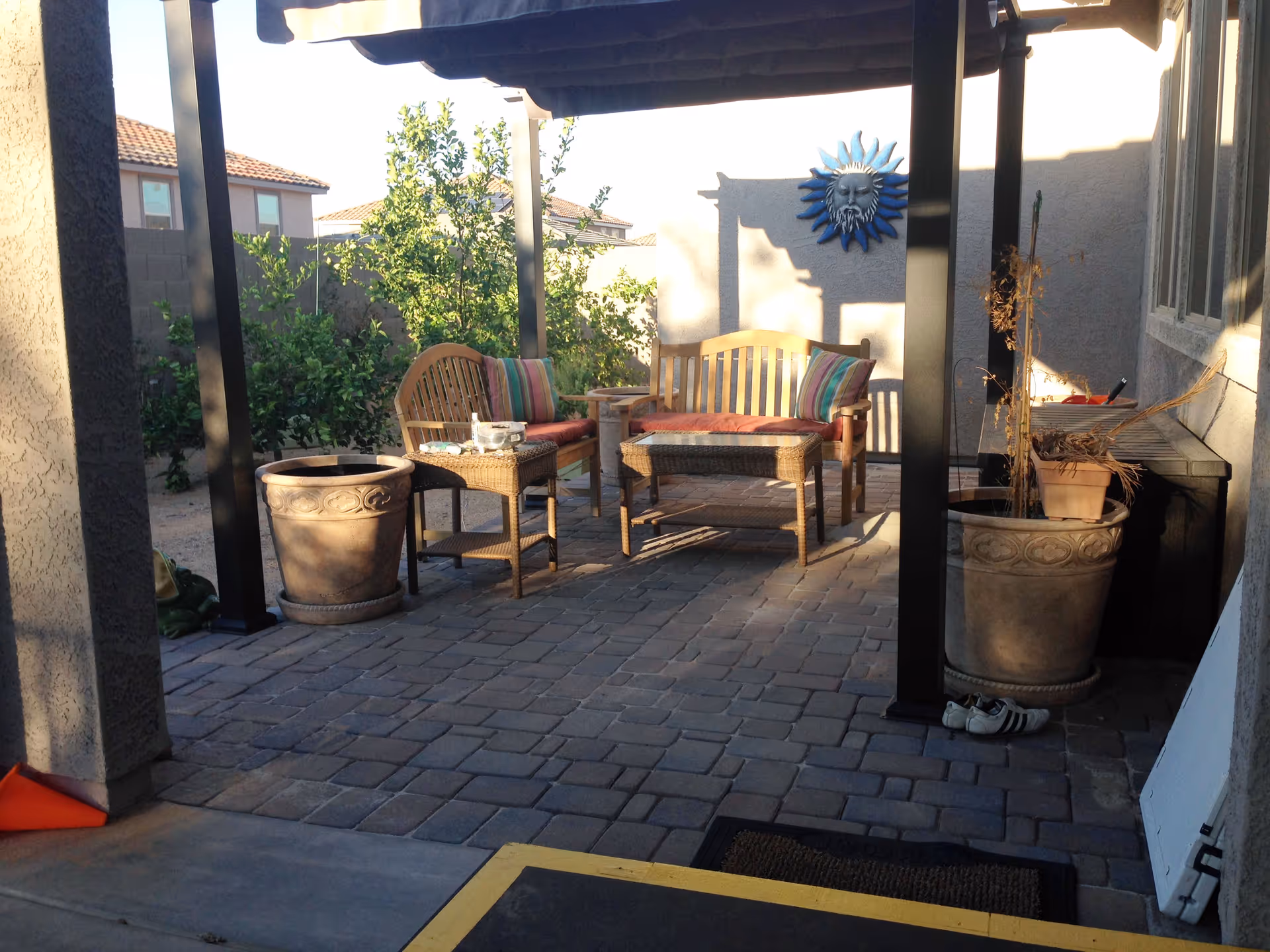 Outdoor patio area with a pergola, two wooden benches with red cushions and striped pillows, a wicker table, large decorative plant pots, and a blue sun face wall decoration. The patio is paved with stone tiles and surrounded by greenery and neighboring houses.