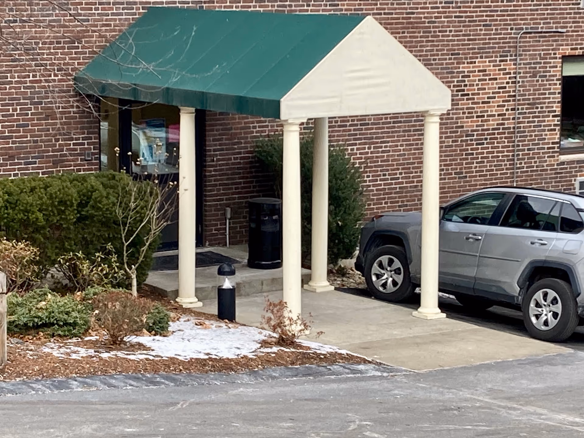 Front entrance of a brick building with a green awning supported by columns and a silver SUV parked nearby.