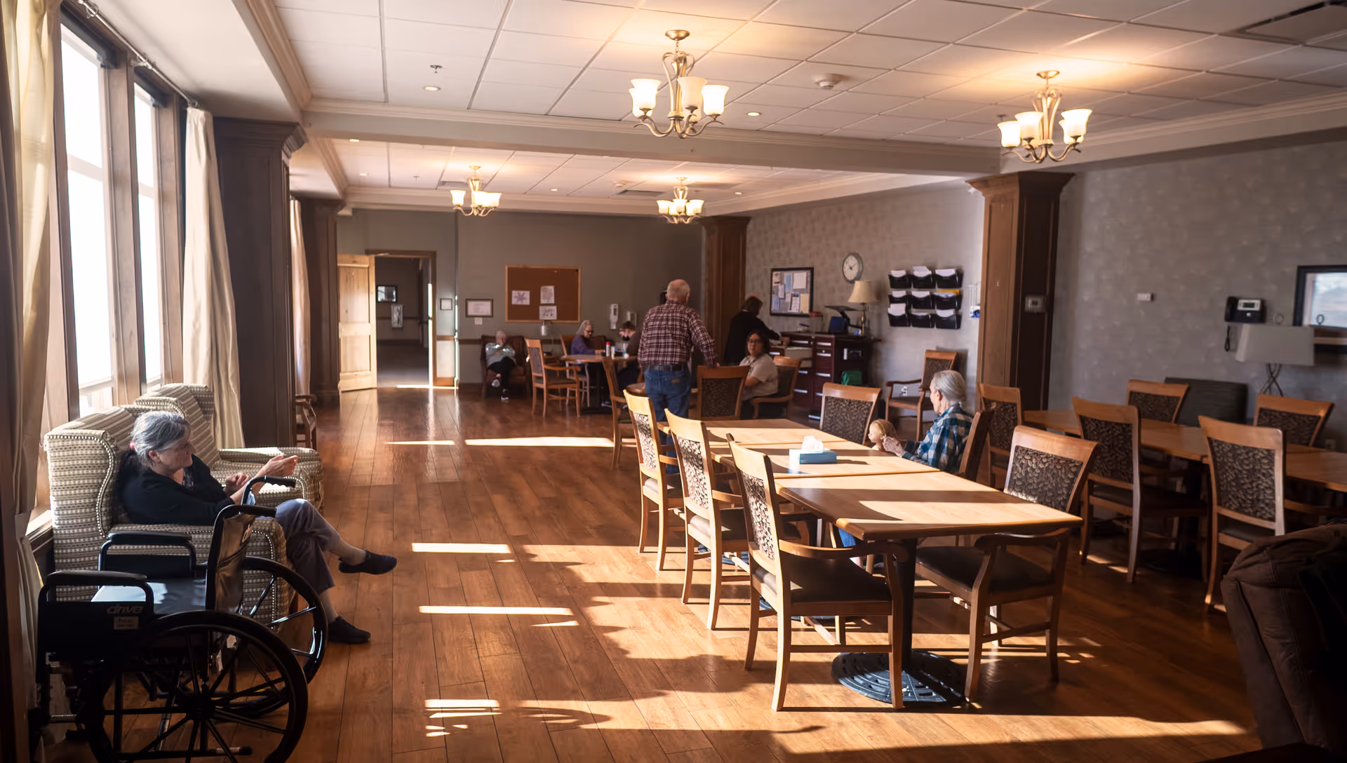 A spacious, well-lit common area in a senior living facility with wooden floors and multiple tables and chairs. Several elderly residents are seated, some in conversation and others relaxing. Large windows on the left side allow natural light to fill the room. The room features chandeliers and a bulletin board on the far wall.