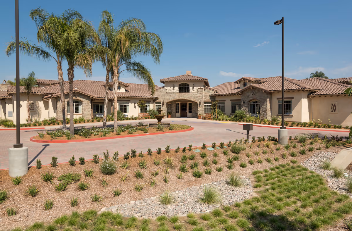 Front exterior view of a single-story senior living facility with a circular driveway, palm trees, landscaped garden beds, and a clear blue sky.
