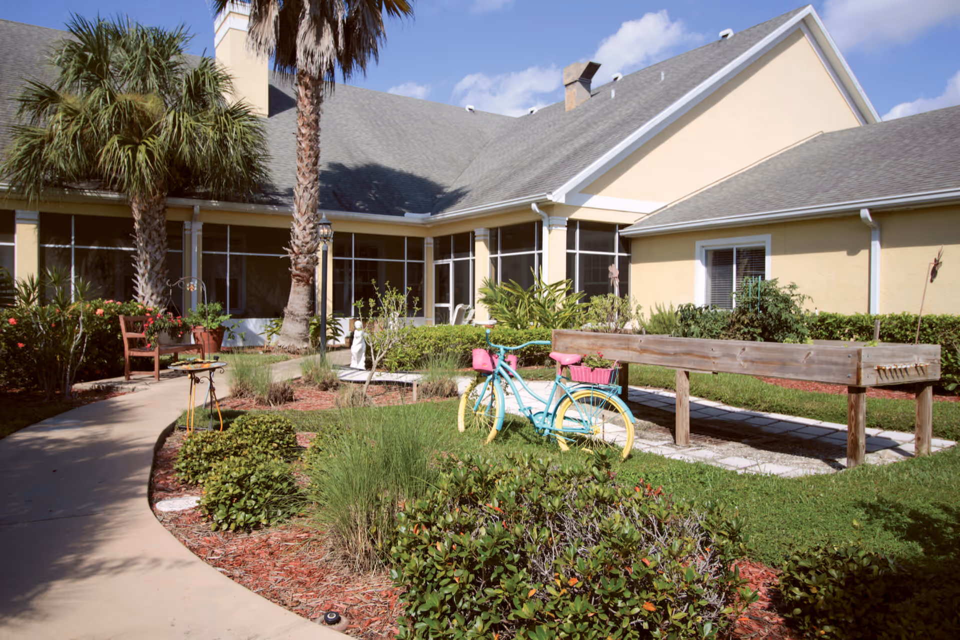 Outdoor garden area at Brookdale Punta Gorda Isles with a curved walkway, palm trees, various plants and shrubs, a decorative blue and yellow bicycle with pink baskets, a wooden raised garden bed, and a yellow building with large windows in the background under a partly cloudy sky.