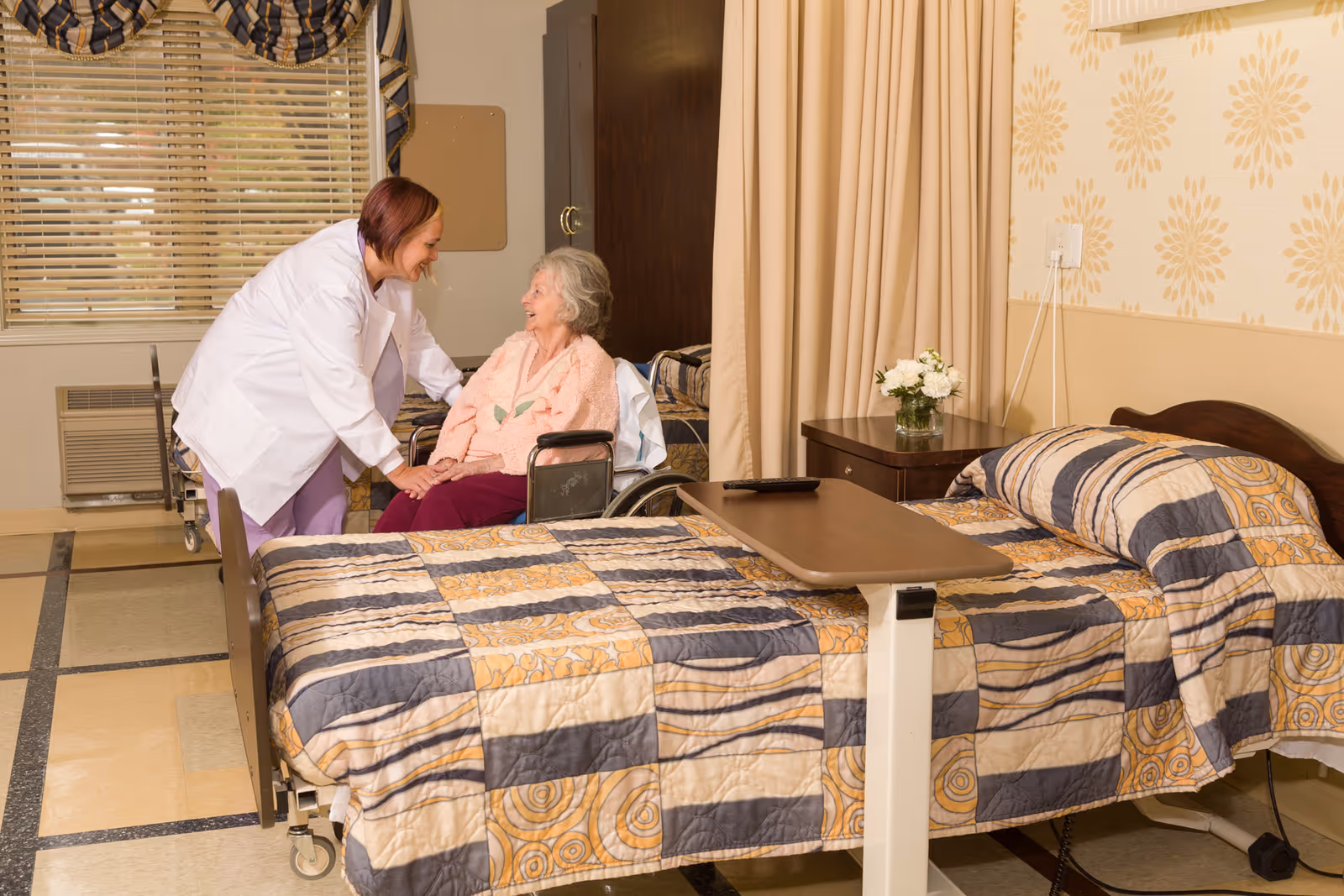 A healthcare worker in a white coat is smiling and holding hands with an elderly woman seated in a wheelchair in a room with a hospital bed covered with a patterned quilt. The room has a window with blinds and a valance, a nightstand with a vase of flowers, and a privacy curtain partially drawn.