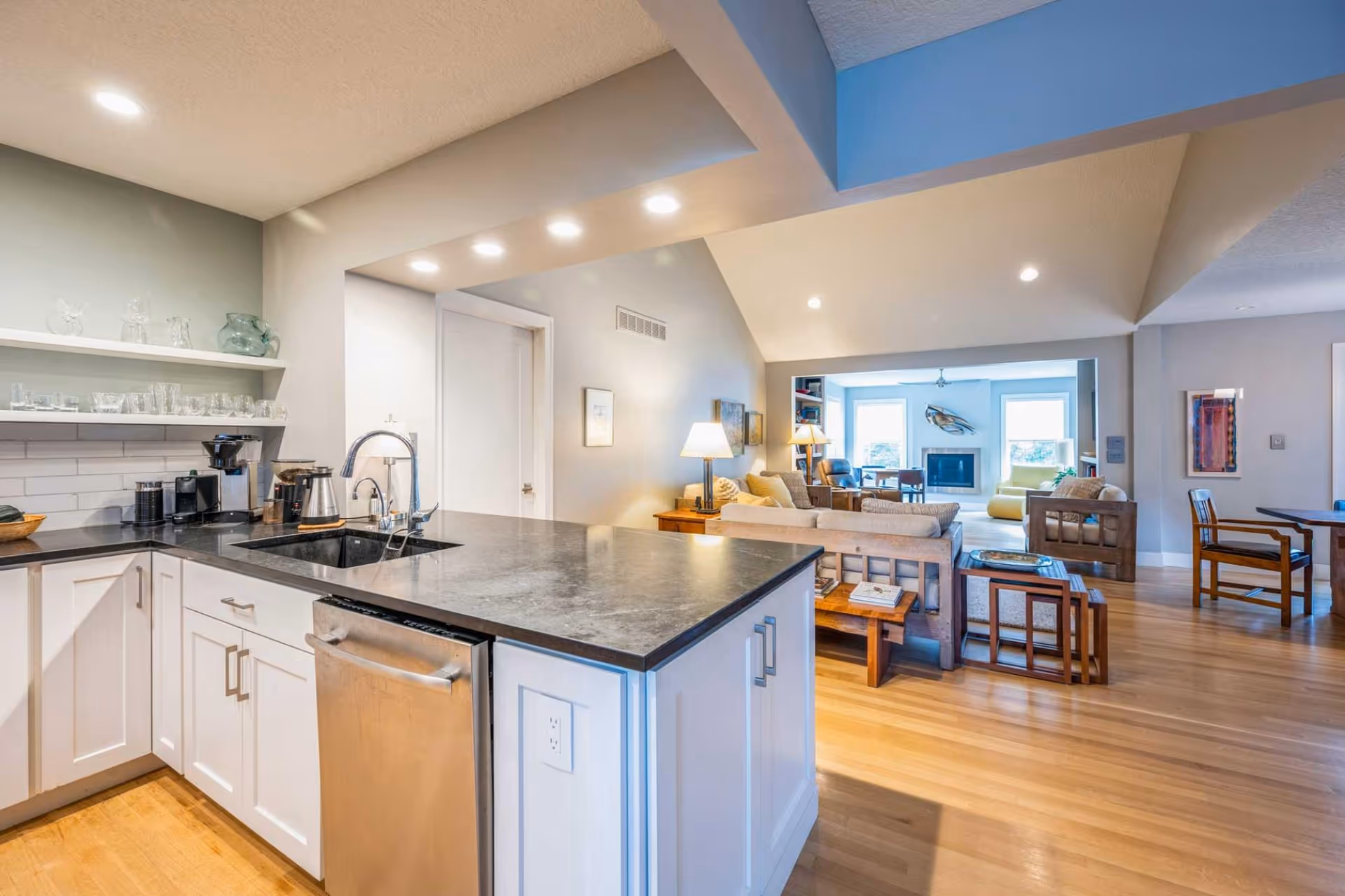 Open-plan kitchen with a large dark countertop island and sink overlooking a bright living room with seating and a fireplace.