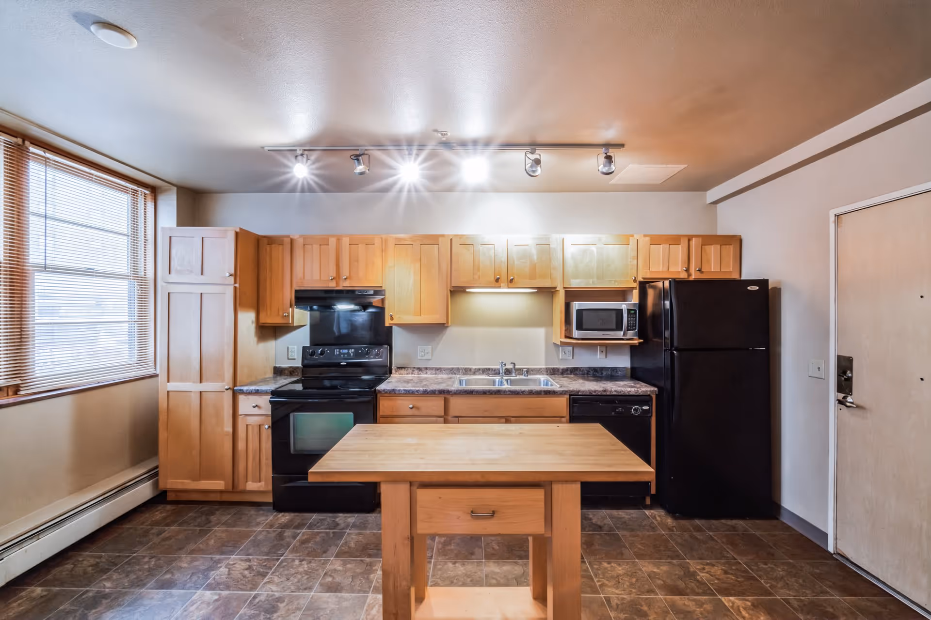 A kitchen area with wooden cabinets, a black stove, black refrigerator, microwave, dishwasher, and a wooden island table in the center. There is a window with blinds on the left side and a door on the right side. The floor is tiled and the ceiling has track lighting.