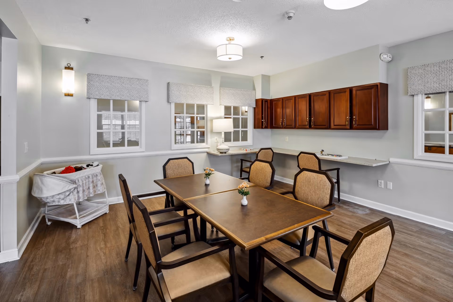 A well-lit room with a rectangular wooden table surrounded by six cushioned chairs. The room has light gray walls, three windows with white frames and patterned valances, and a wooden floor. On the left side, there is a white bassinet with a blanket and stuffed animals. On the right wall, there are dark wooden cabinets mounted above a long white countertop with two chairs underneath. A table lamp is placed on the countertop near the windows.