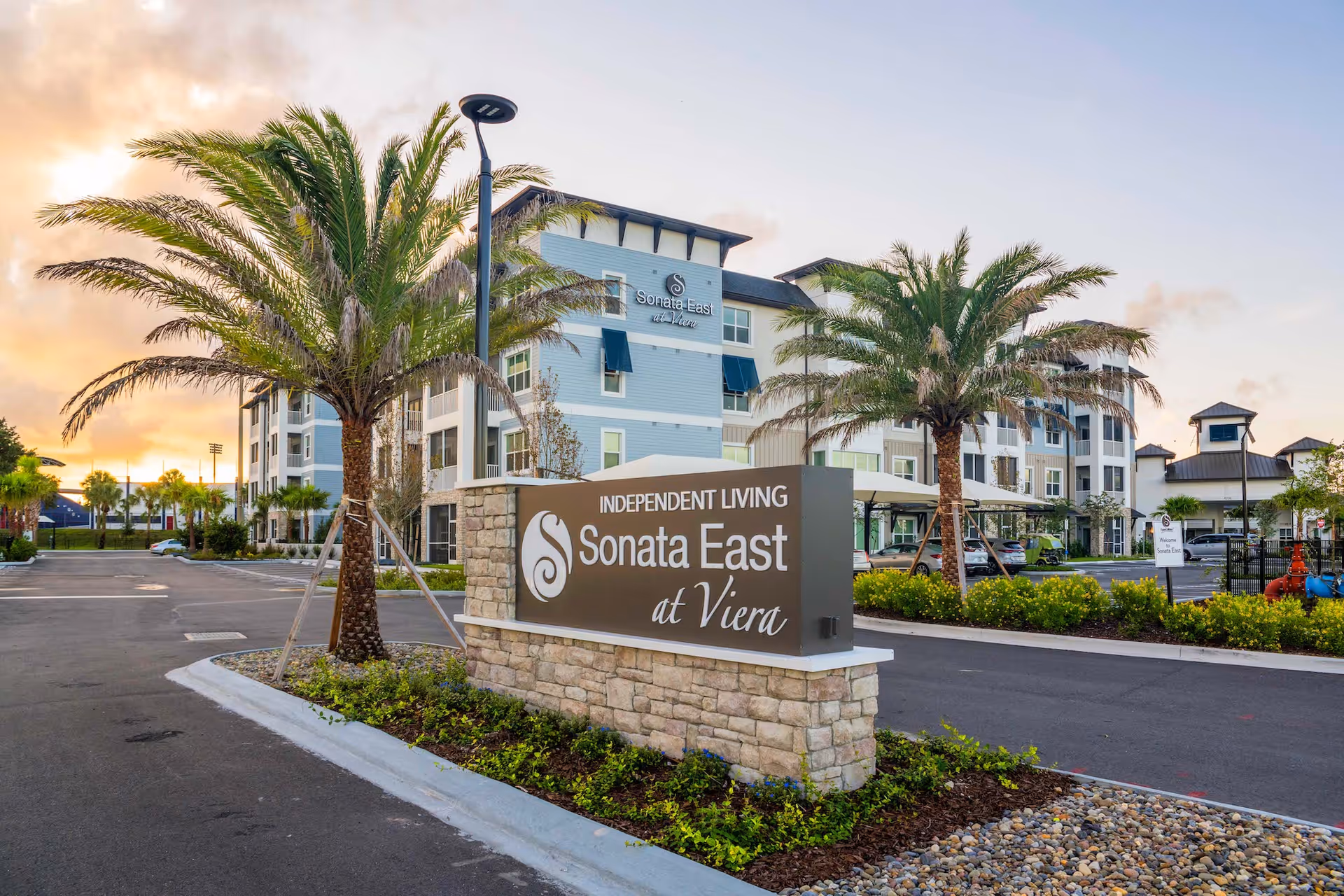 Exterior view of Sonata East at Viera senior living facility during sunset, showing a large sign that reads 'Independent Living Sonata East at Viera' in front of a multi-story building with palm trees and a parking area.