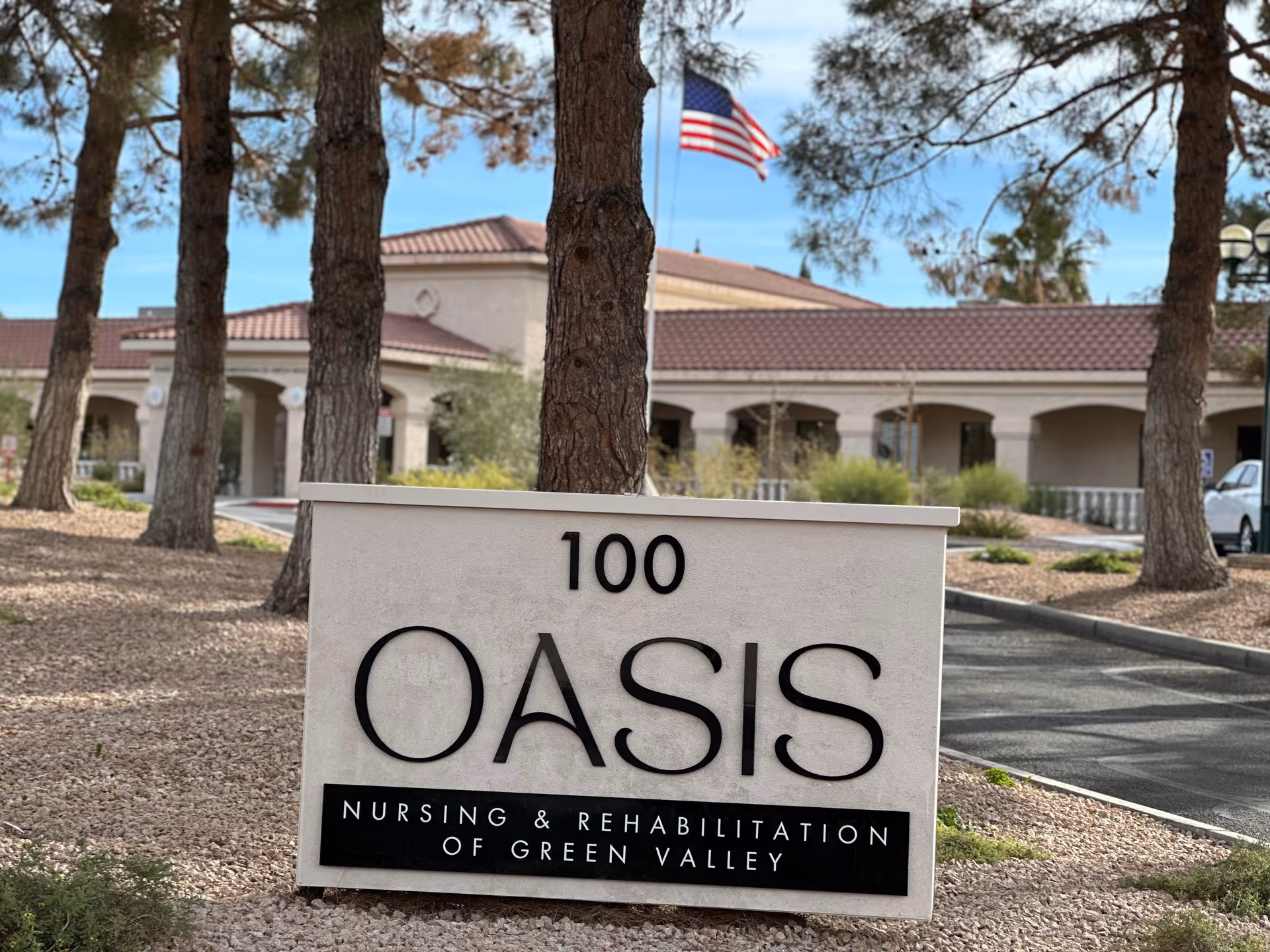 Outdoor view of the entrance area of Oasis Nursing & Rehabilitation of Green Valley, featuring a large sign with the facility name and address number 100 in front of several trees and a building with a tiled roof. An American flag is visible flying in the background.