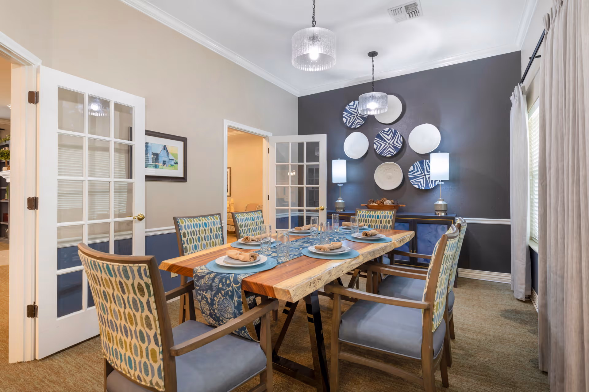 A dining room with a wooden table set for six people, featuring blue and beige patterned chairs. The table has a blue runner, plates, napkins, and glasses. The room has a dark accent wall decorated with circular wall art and two table lamps on a sideboard. French doors and a window with curtains are also visible.