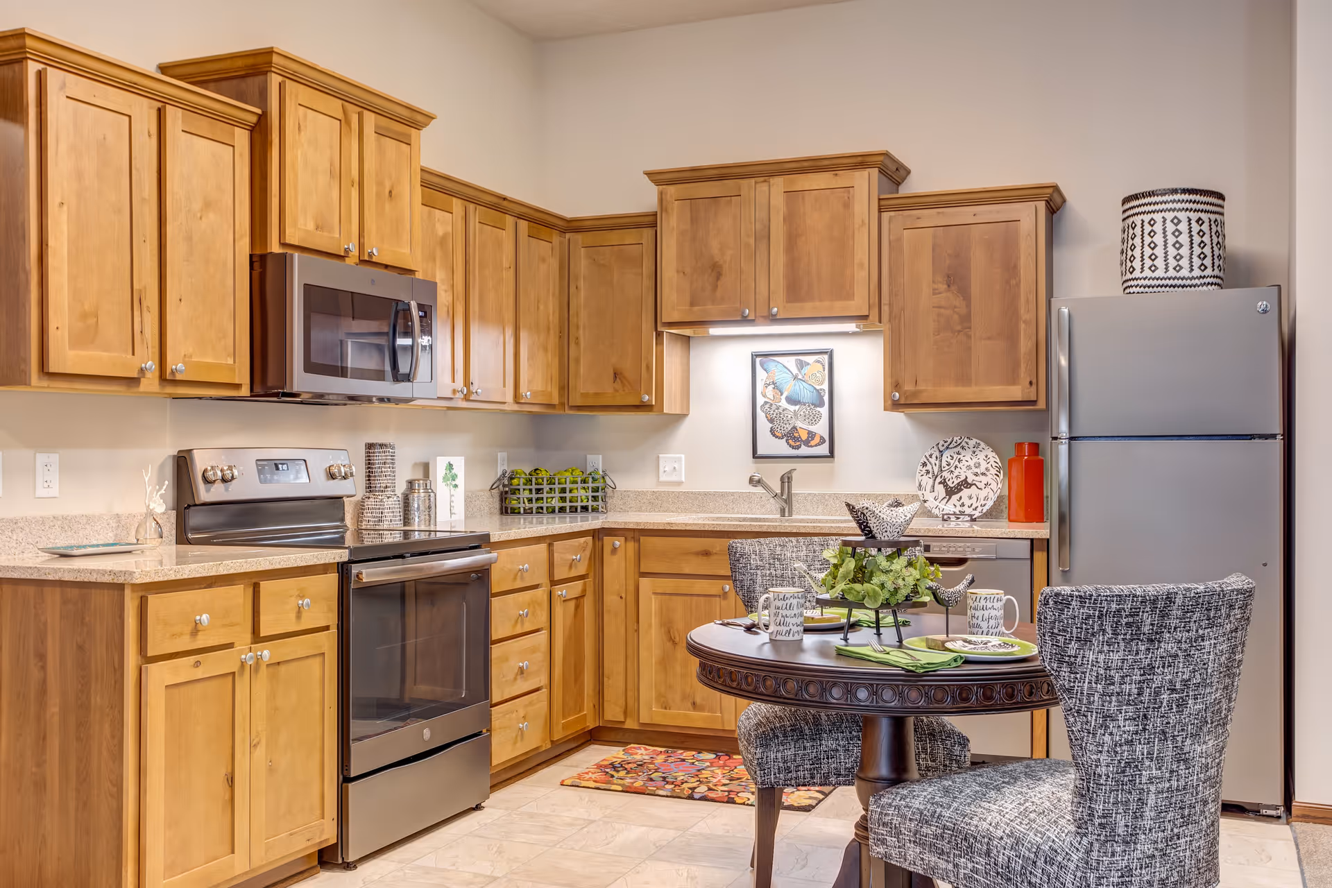 A modern kitchen with wooden cabinets, stainless steel appliances including a microwave, stove, and refrigerator. There is a small round dining table with two upholstered chairs, set with mugs, plates, and a decorative centerpiece. The kitchen has a light-colored countertop and a framed butterfly artwork on the wall above the sink.
