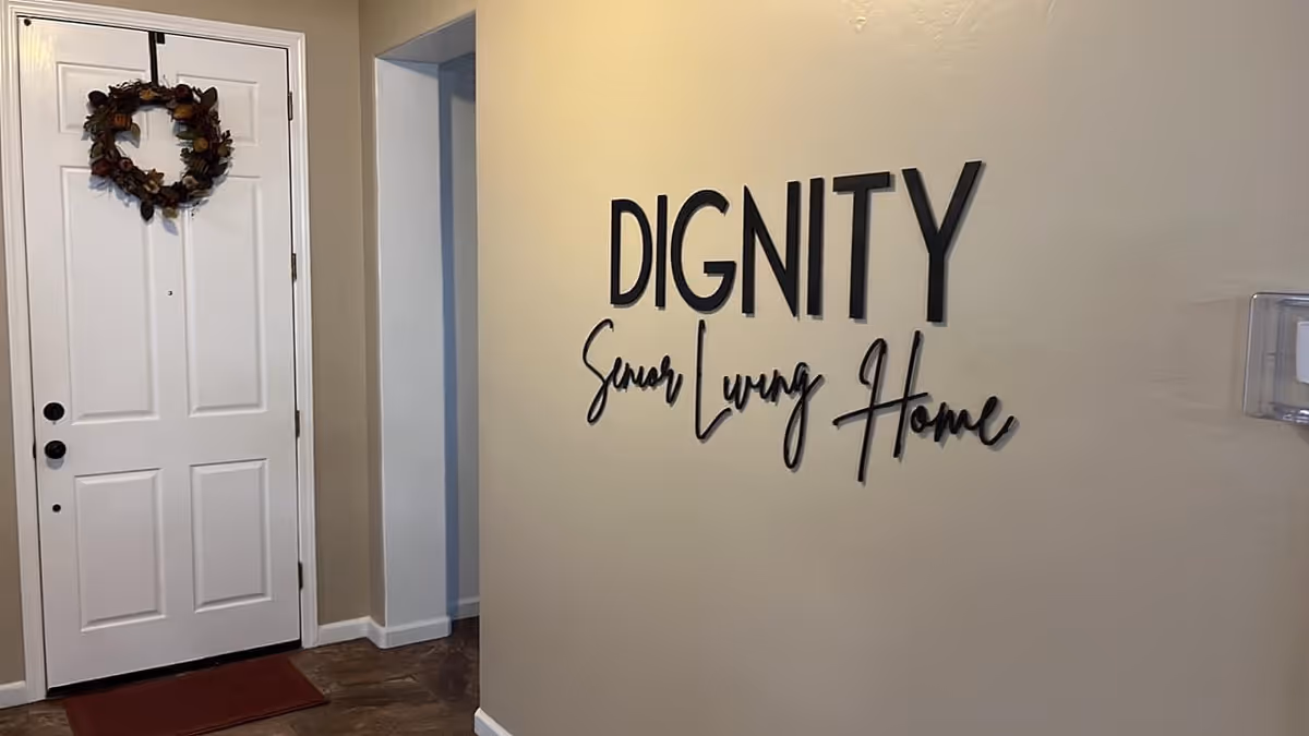Interior view of an entryway with a white door decorated with a wreath on the left and a wall on the right displaying the text 'DIGNITY Senior Living Home' in black letters.