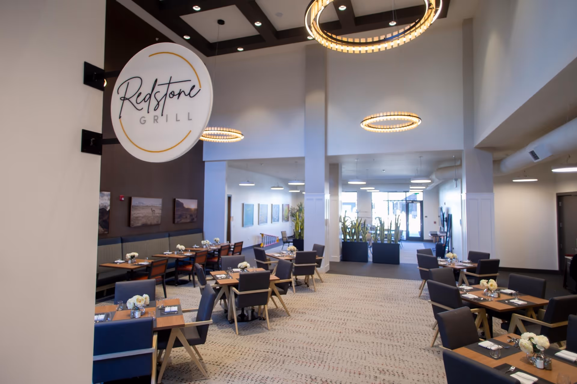 Interior view of a modern dining area named Redstone Grill with several tables set with white flowers, glasses, and napkins. The space features high ceilings with circular hanging lights, cushioned chairs, and large windows letting in natural light. There are plants and artwork on the walls, creating a welcoming atmosphere.