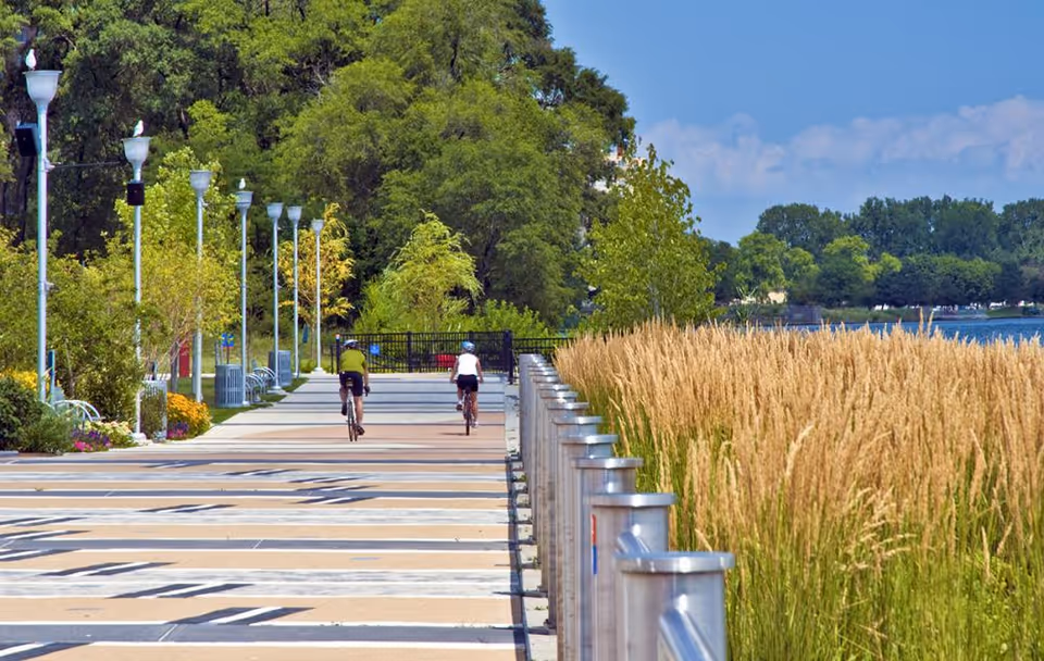 Two people riding bicycles on a paved pathway lined with tall grass on one side and trees and lampposts on the other, with a body of water visible in the background under a partly cloudy sky.