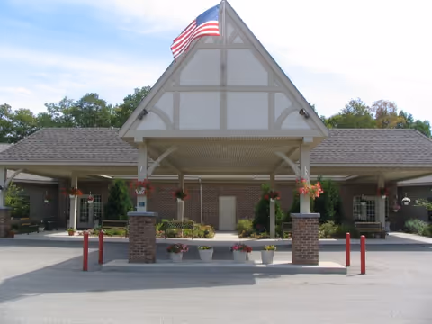 Front exterior view of a retirement village building with a peaked roof, an American flag on top, brick pillars, hanging flower baskets, and potted plants near the entrance.
