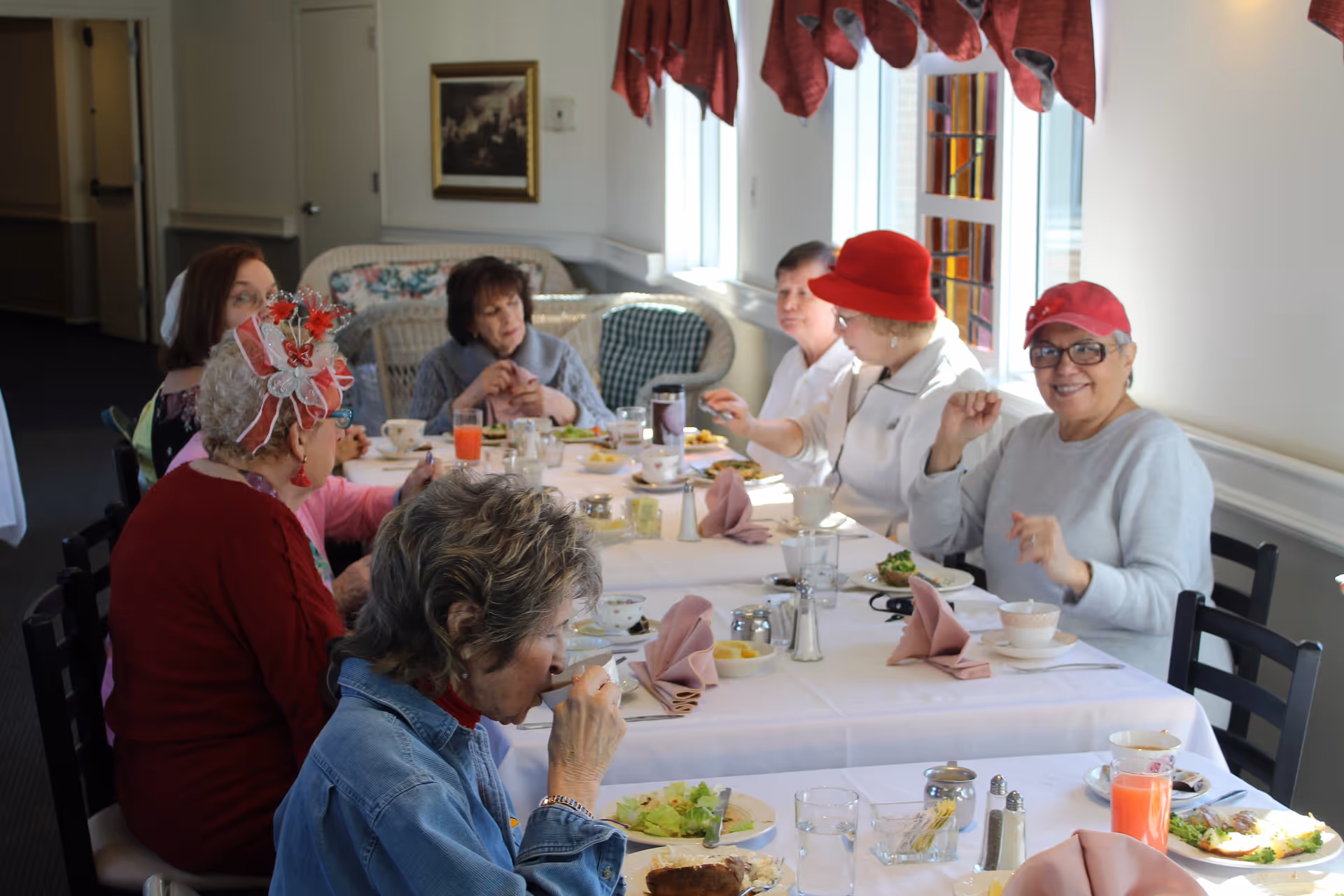 A group of elderly women sitting around a dining table enjoying a meal together in a well-lit room with windows and red curtains. The table is set with plates of food, glasses, cups, and folded napkins.