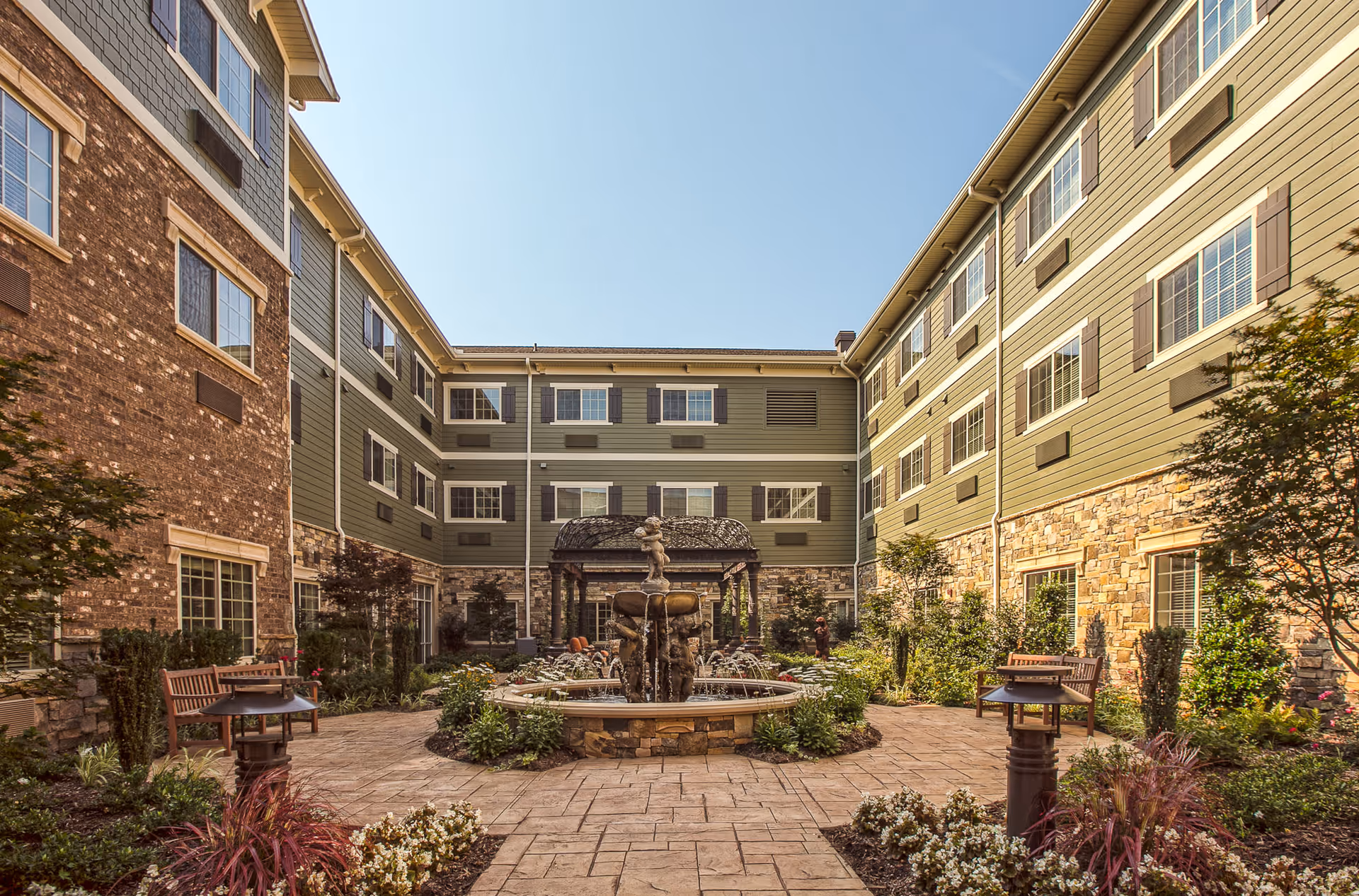 Enclosed sunlit courtyard with a central fountain, benches, landscaping and surrounding multi-story senior living building.
