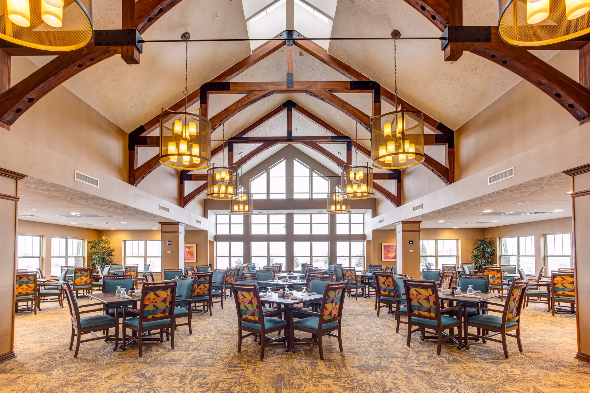 Spacious dining room with high vaulted ceilings featuring exposed wooden beams and large hanging light fixtures. Multiple tables and chairs with colorful patterned backs are arranged neatly across the carpeted floor. Large windows at the far end allow natural light to fill the room, and there are plants and framed artwork on the walls.