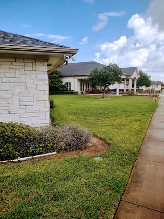 Front exterior of a single-story building with white stone, columns, a manicured lawn and a sidewalk under a partly cloudy sky.