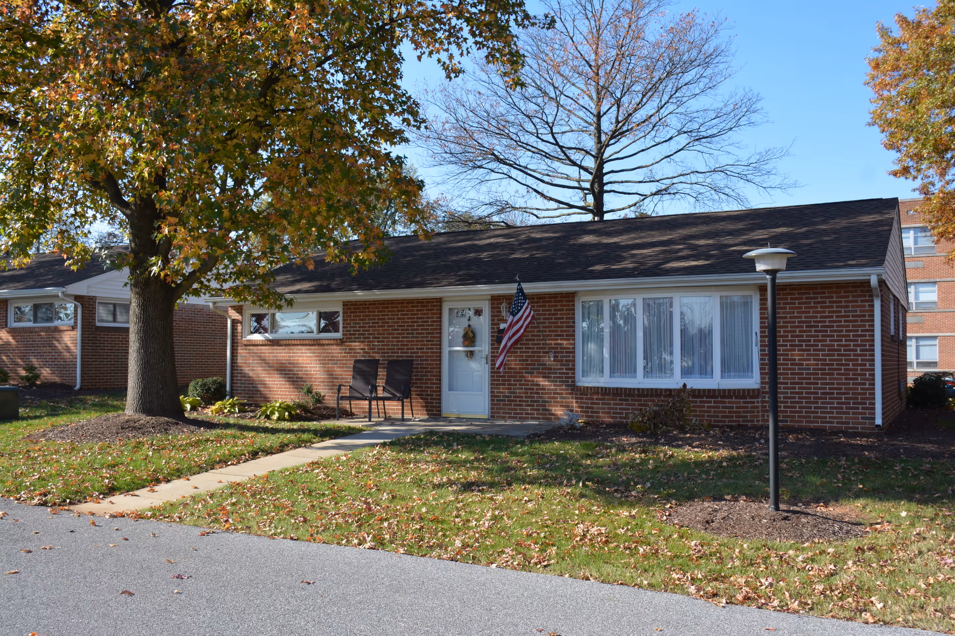 Single-story brick building with a white door and large window, two chairs on the front porch, an American flag hanging beside the door, a tree with autumn leaves in the front yard, and a lamp post near the sidewalk.
