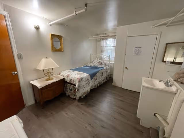 Small bedroom in a senior living facility with bed, floral cover, nightstand, lamp, sink, and door.