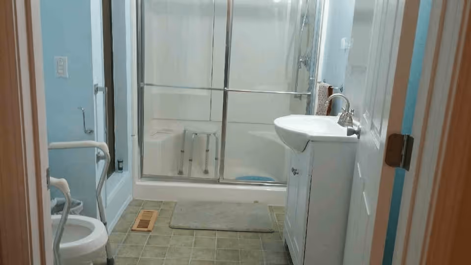 A bathroom with a white toilet equipped with safety rails, a white vanity with a sink and faucet, a glass-enclosed shower with a white shower stool inside, a beige bath mat on the tiled floor, and light blue walls.