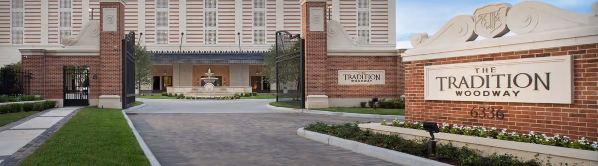 Entrance to The Tradition Woodway senior living facility featuring a brick driveway, gated entry, manicured lawns, and a large sign with the facility name and address. The building is visible in the background with a fountain in front.