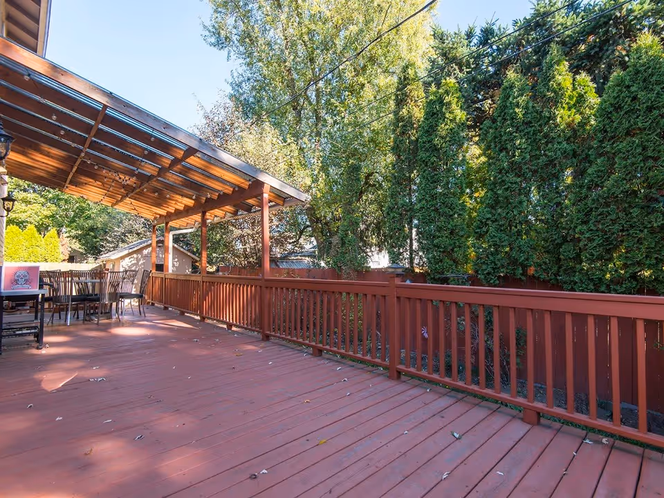 A spacious outdoor wooden deck with a red railing and a covered seating area with chairs and a table. The deck is surrounded by tall green trees and bushes under a clear blue sky.