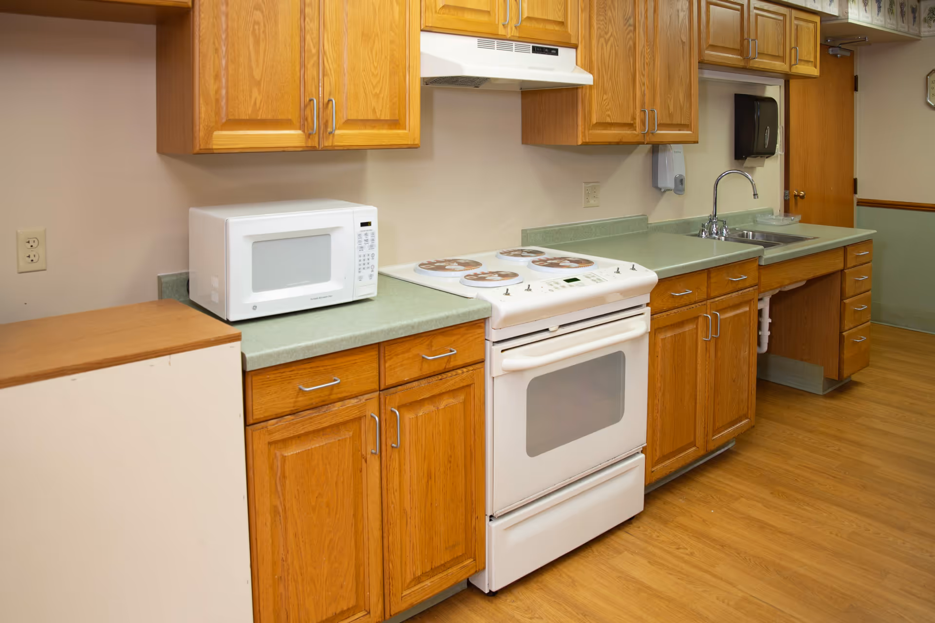 A kitchen area with wooden cabinets, a white microwave on the countertop, a white electric stove with four burners, a stainless steel sink with a faucet, and a paper towel dispenser mounted on the wall. The floor is wooden, and there is a door in the background.