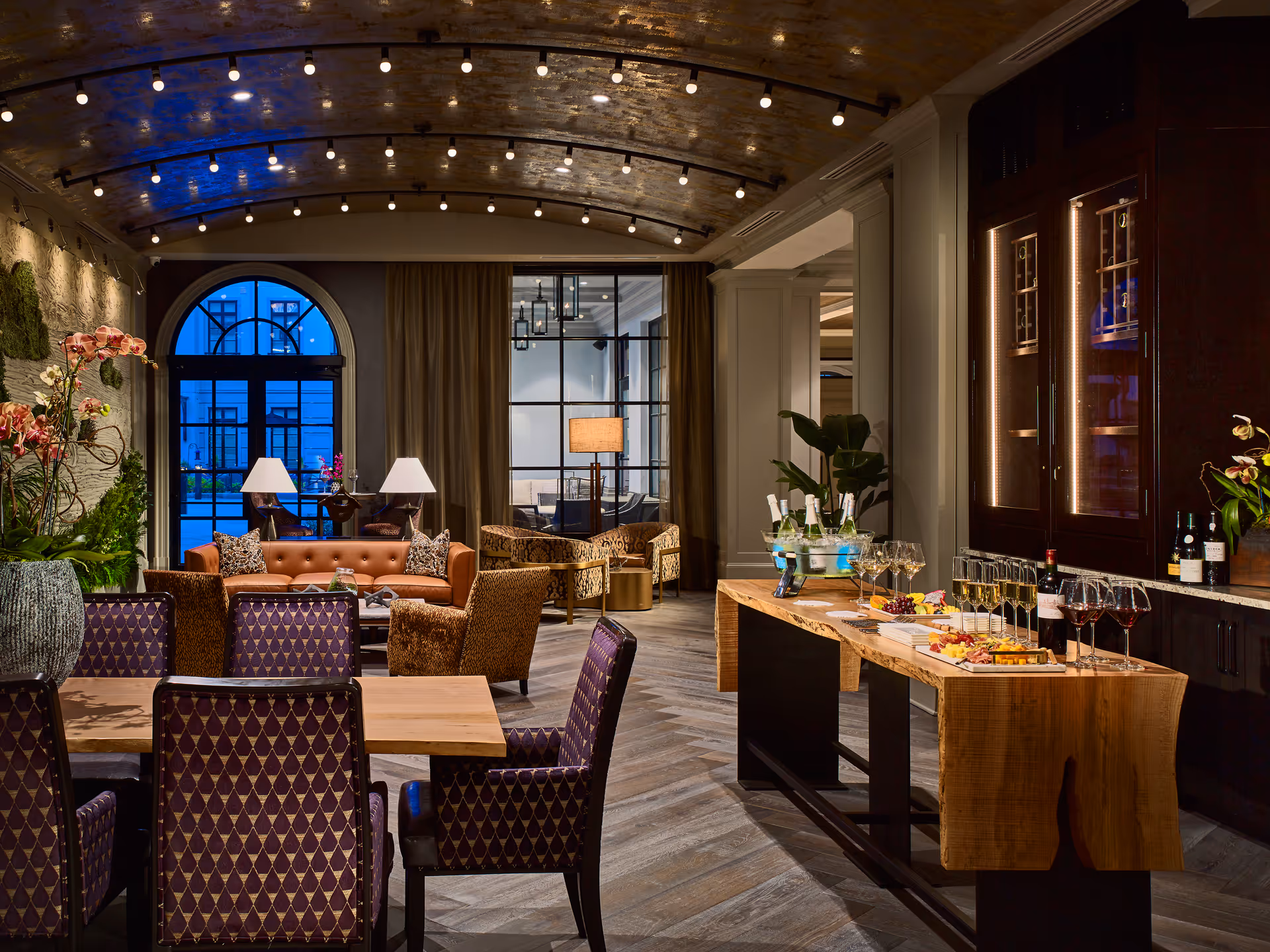 A stylish and warmly lit lounge area featuring a wooden table set with wine glasses, bottles, and appetizers. The room has a curved ceiling with exposed brick and string lights, a large arched window with blue light outside, comfortable seating including a brown leather sofa and patterned chairs, and decorative plants.