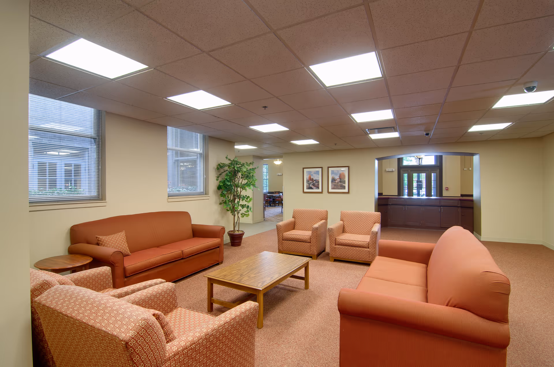 A well-lit senior living facility common area with two orange sofas, two patterned armchairs, a wooden coffee table, a side table, and a potted plant. The room has beige walls, carpeted floor, and several windows with blinds. Two framed pictures hang on the wall, and there is an open doorway leading to another room with more seating and a reception area visible in the background.