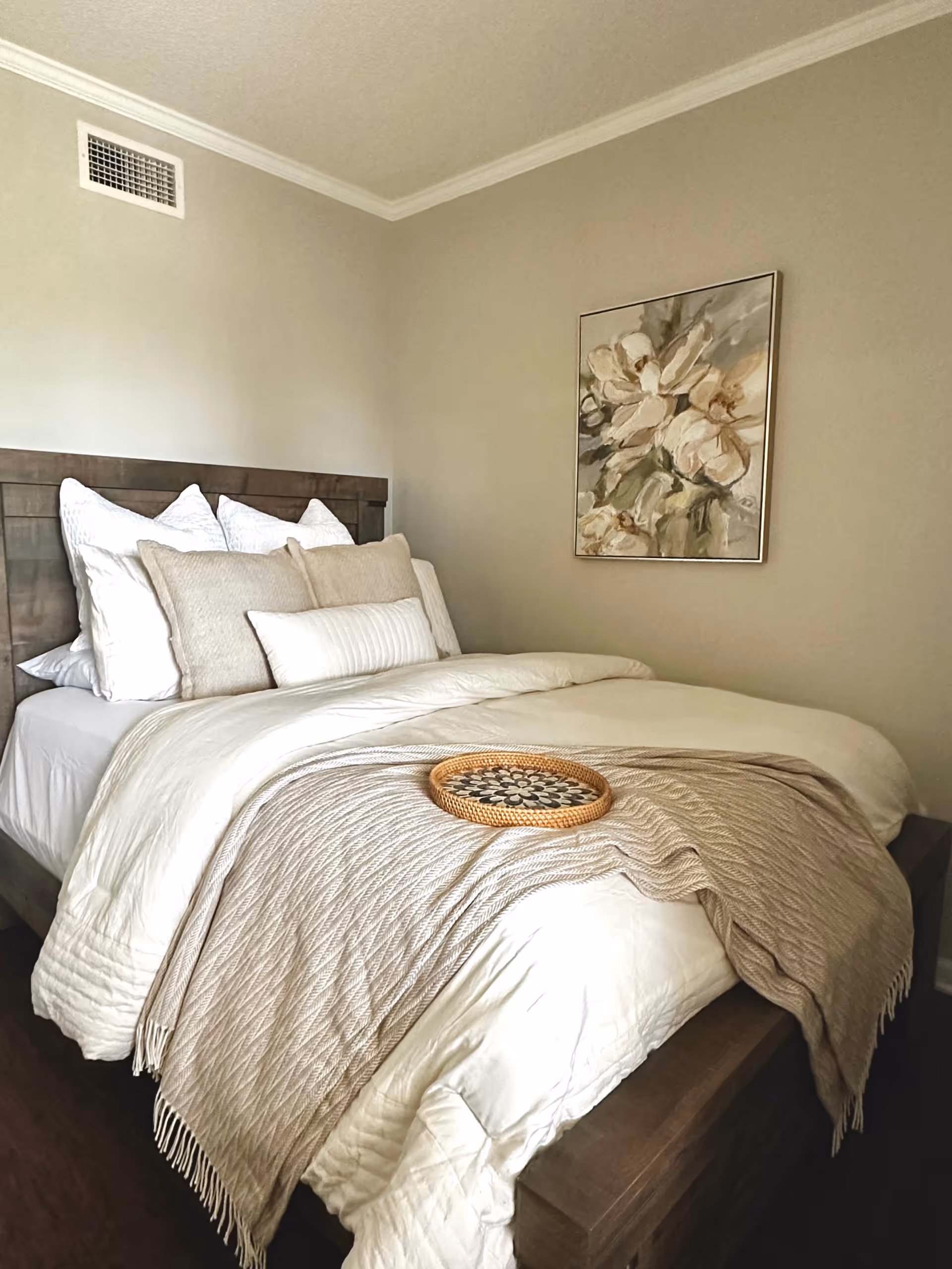 A neatly made bed with multiple pillows and a beige knitted throw blanket draped over the foot of the bed. A decorative round tray is placed on the blanket. The room has beige walls with a framed floral painting hanging above the bed's wooden headboard.