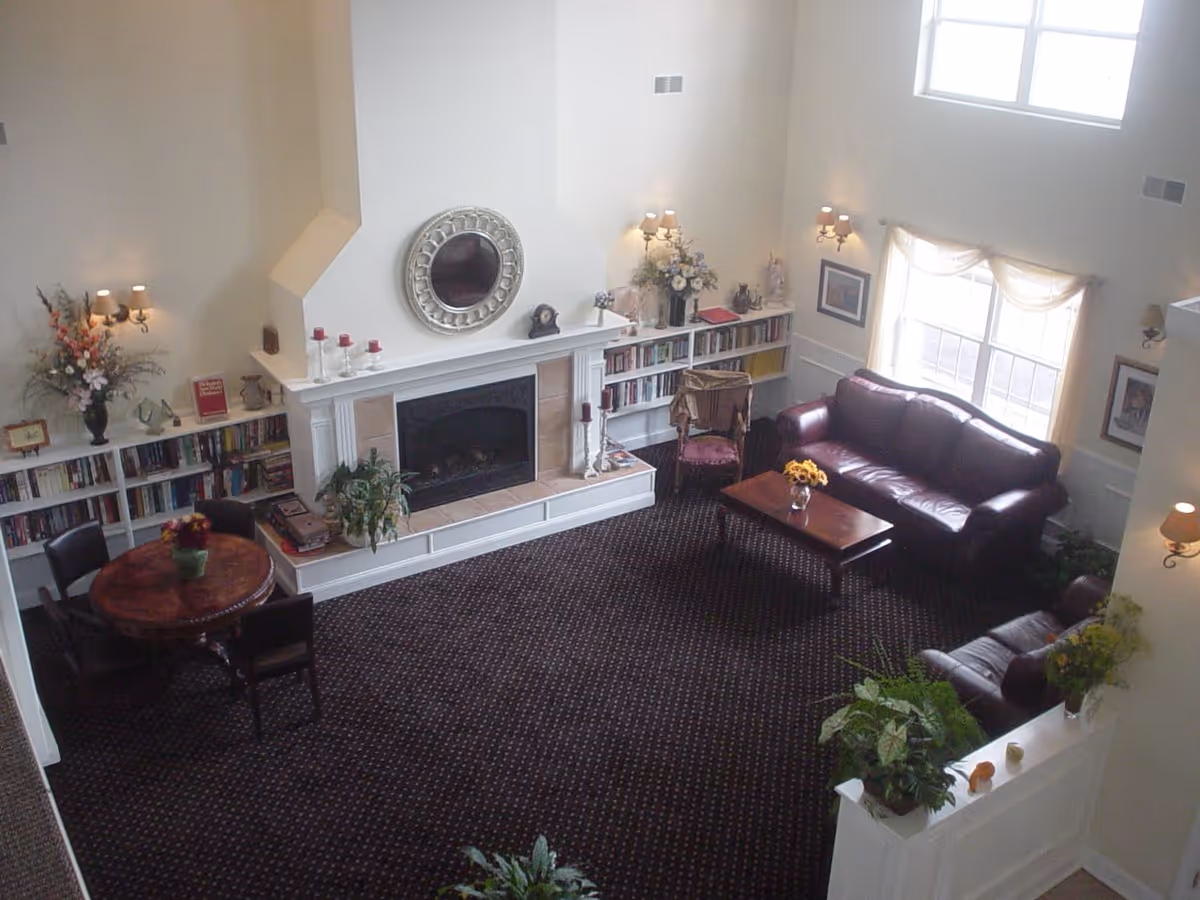 A cozy living room with a dark patterned carpet, a white fireplace with a round decorative mirror above it, and built-in bookshelves on either side. The room features a round wooden table with four chairs, two leather sofas, a wooden coffee table with a vase of flowers, and several plants and floral arrangements. Large windows with sheer curtains allow natural light to fill the space.