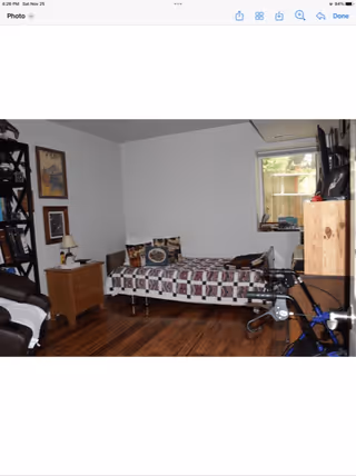 A cozy bedroom with a single bed covered in a patterned quilt and decorative pillows. There is a wooden nightstand with a lamp and some items on it next to the bed. A black bookshelf filled with books and decorative items is on the left side of the room. The floor is wooden, and there is a window with a view of an outdoor area. A walker is partially visible in the foreground on the right side.