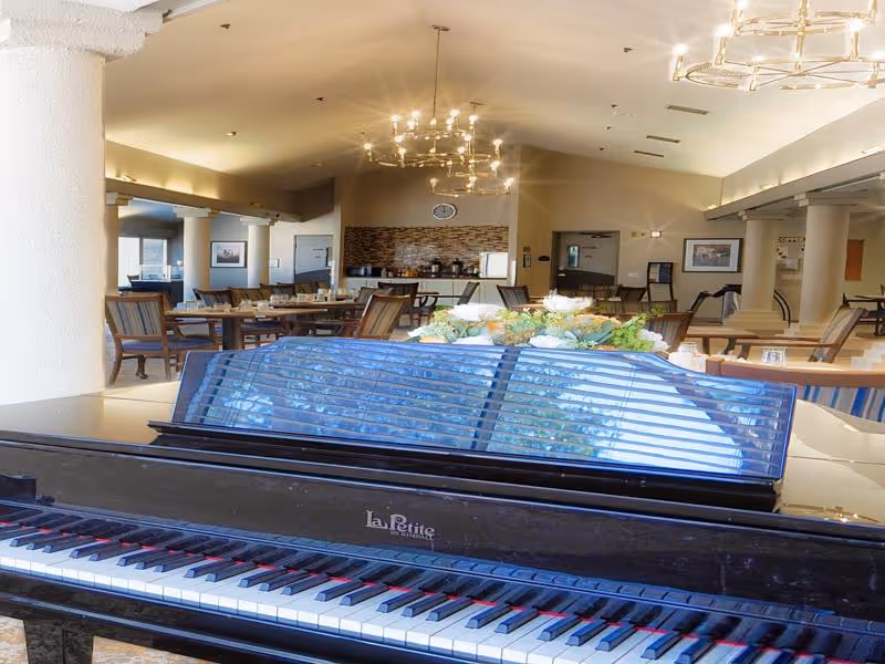 Interior view of a senior living facility common area featuring a black La Petite piano in the foreground, with several tables and chairs arranged in the background under warm lighting and chandeliers.