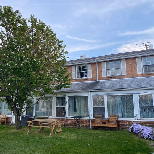 Outdoor view of a two-story brick building with white framed windows and a gray roof. In front of the building is a green lawn with a large leafy tree, wooden picnic tables, benches, and a small patch of purple flowers. The sky is partly cloudy.