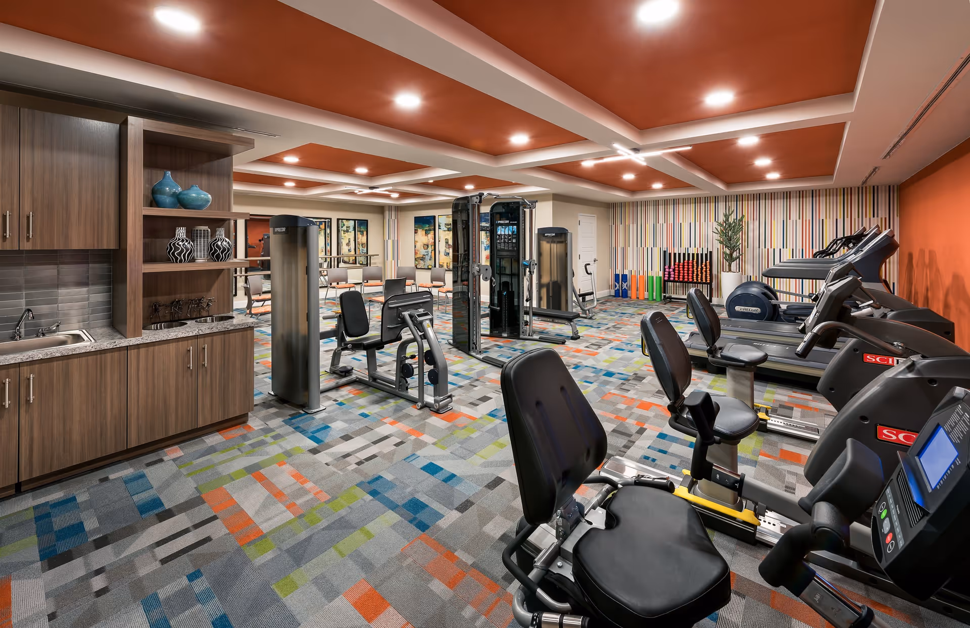 A brightly lit fitness room with various exercise equipment including recumbent bikes, treadmills, and strength training machines. The room features a colorful patterned carpet, orange ceiling panels with recessed lighting, and a wall with vertical striped wallpaper. There is a wooden cabinet with a sink and decorative vases on shelves to the left, and chairs arranged along the back wall.
