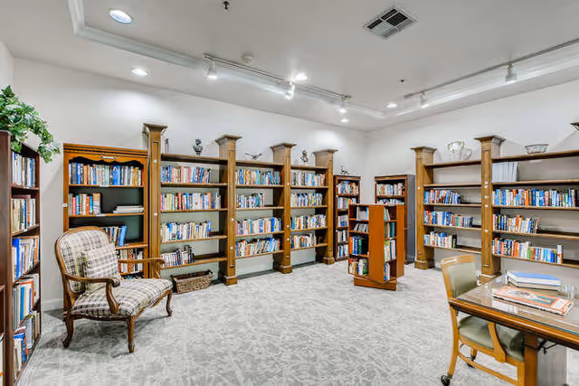 A well-lit library room with multiple wooden bookshelves filled with books along the walls and a few freestanding shelves in the center. There is a cushioned armchair with a plaid pattern on the left side and a wooden table with chairs on the right side. The room has a light carpeted floor and white walls with ceiling lights.