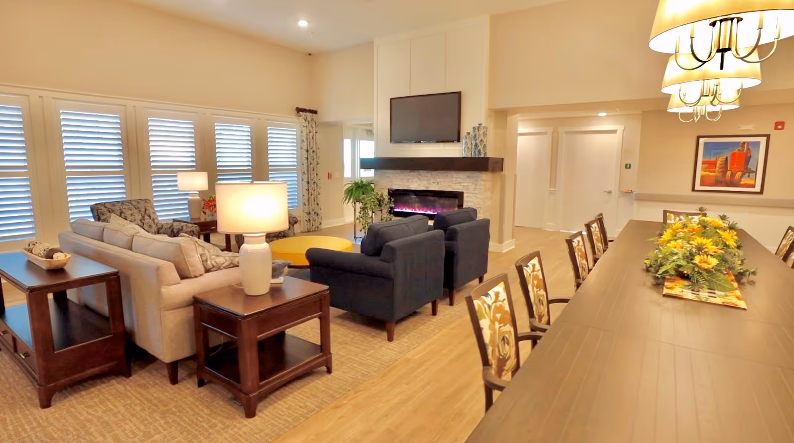 Bright communal living area with sofas and armchairs arranged around a fireplace and a long dining table with a floral centerpiece.