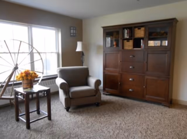 A cozy living room area with a large window letting in natural light. There is a comfortable armchair next to a small wooden side table with a flower arrangement on it. Behind the chair is a tall floor lamp, and against the wall is a large wooden cabinet with shelves and drawers containing books and baskets.