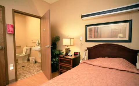 A bedroom with a nightstand, lamp, framed art, and an open door to a bathroom.