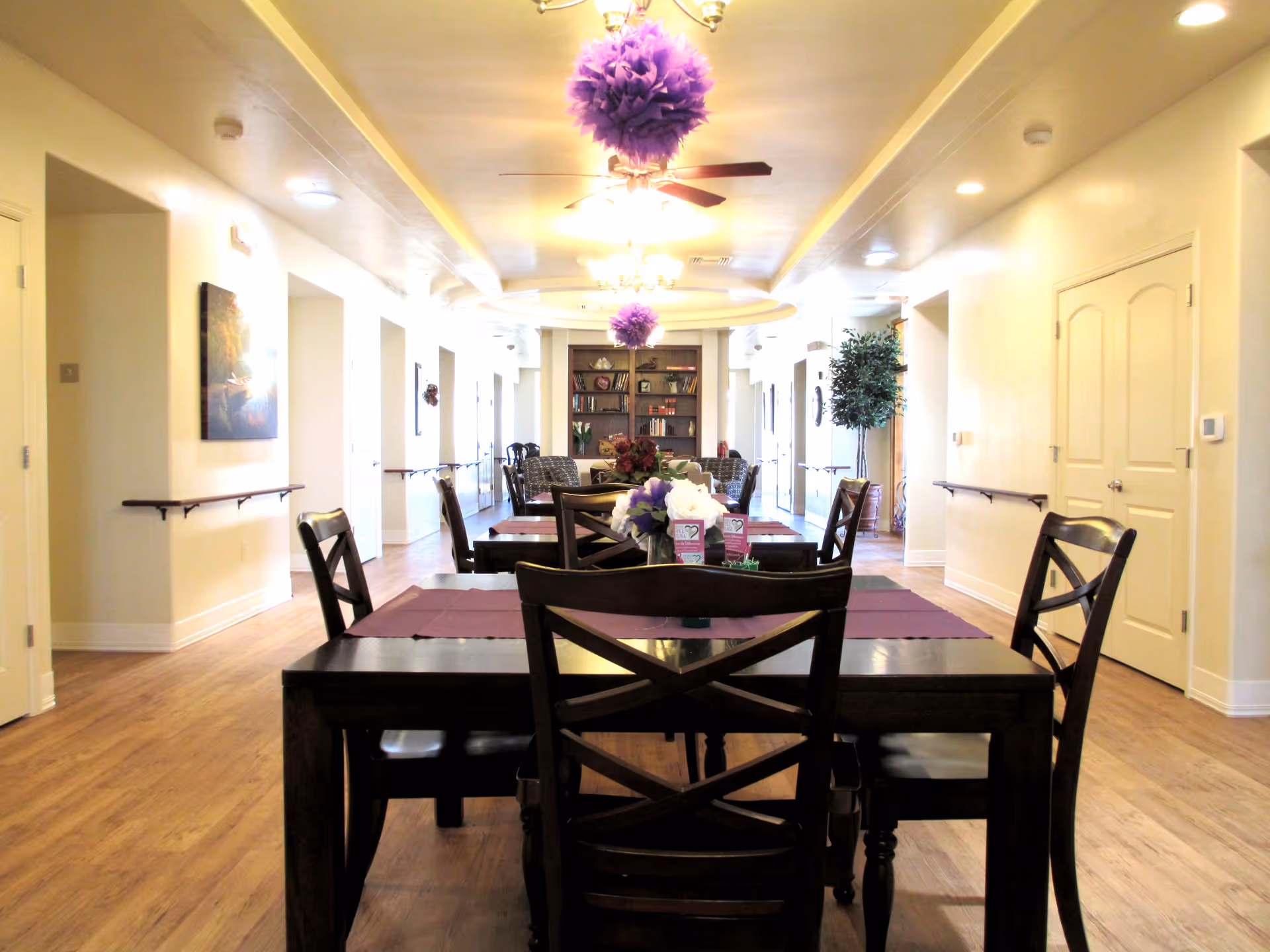 Interior view of a senior living facility dining area with wooden tables and chairs arranged in rows. The room has light-colored walls, wooden flooring, ceiling fans with lights, and purple decorative hanging pom-poms. There are handrails along the walls and a bookshelf with various items at the far end of the room.